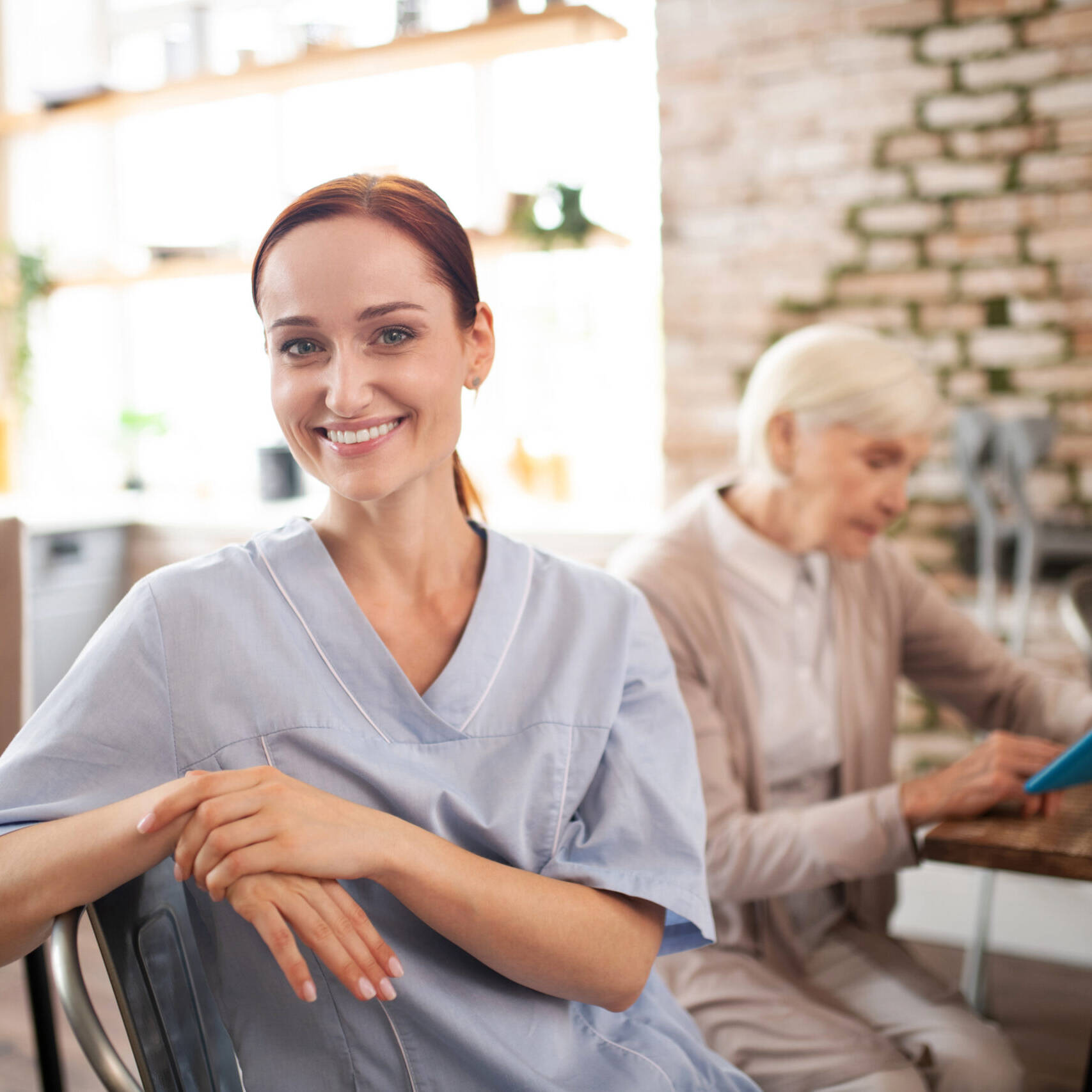 Smiling caregiver in a blue uniform assisting an elderly woman at home. Home care jobs in Philadelphia offer competitive pay and benefits.