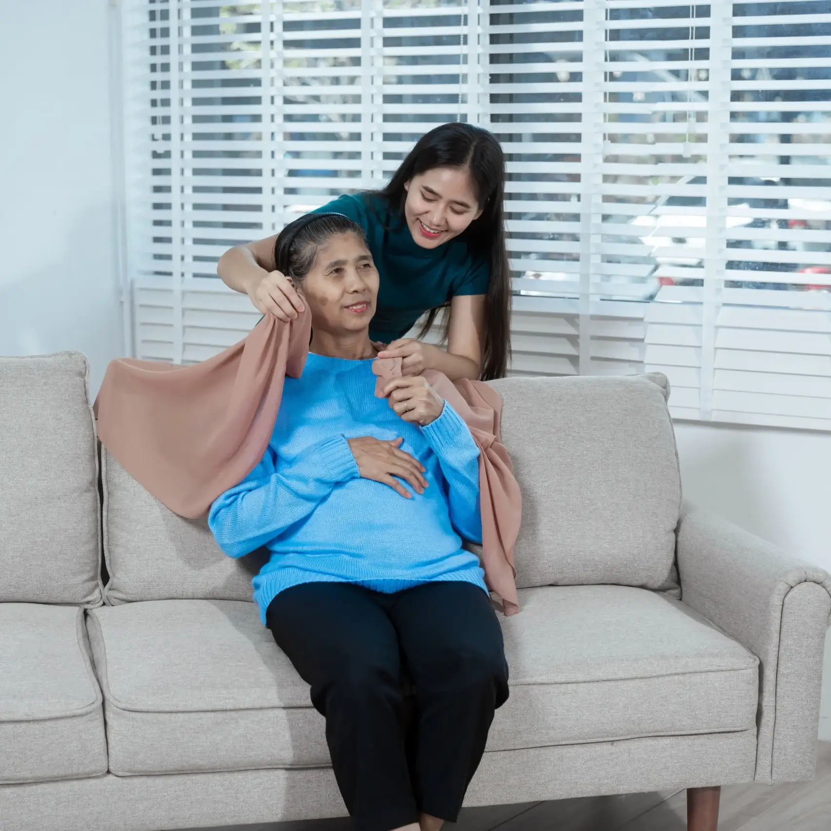 A young woman helps her elderly mother put on a shawl while sitting together on a sofa at home, representing compassionate home caregiving.