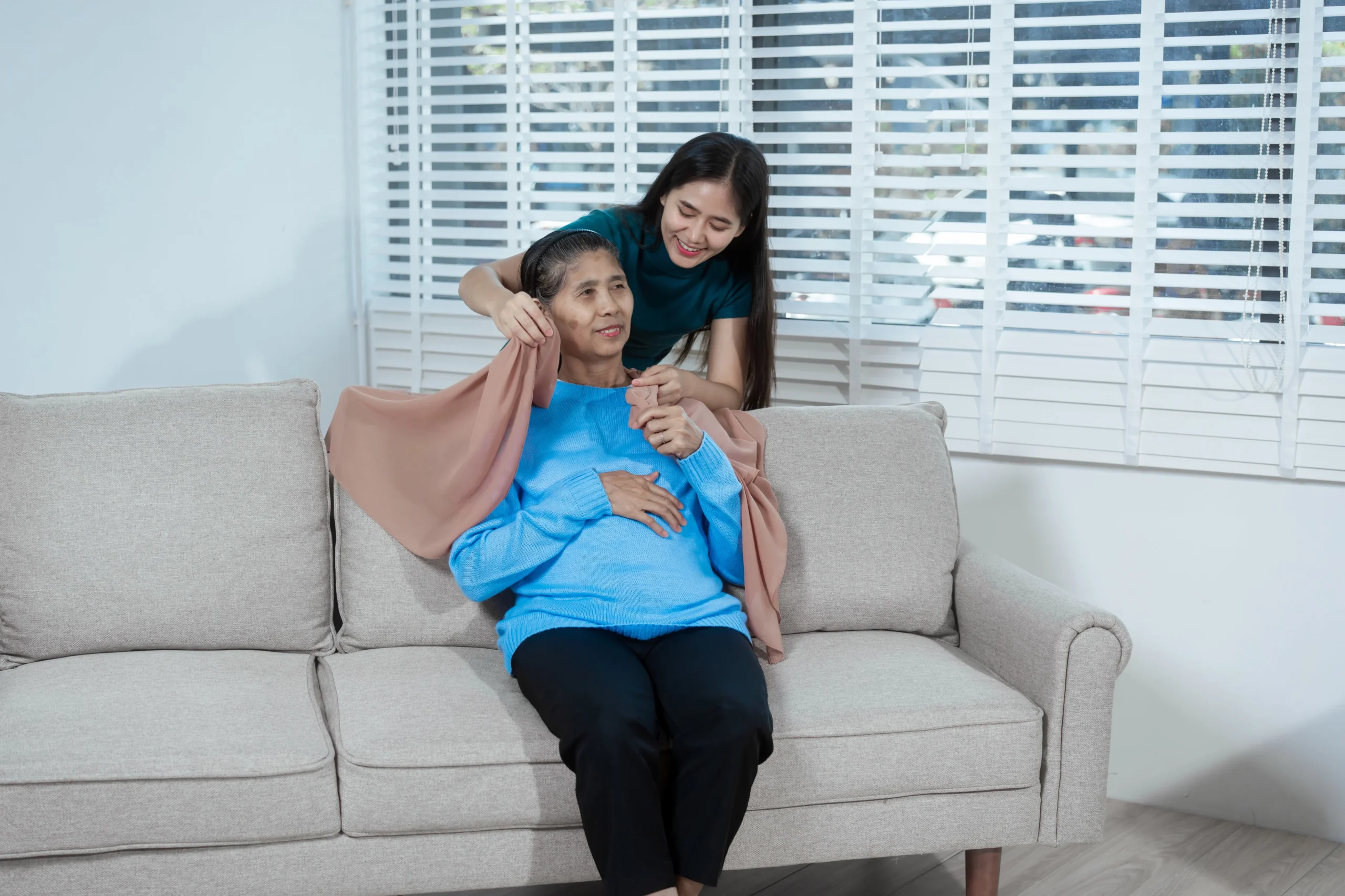 A young woman helps her elderly mother put on a shawl while sitting together on a sofa at home, representing compassionate home caregiving.