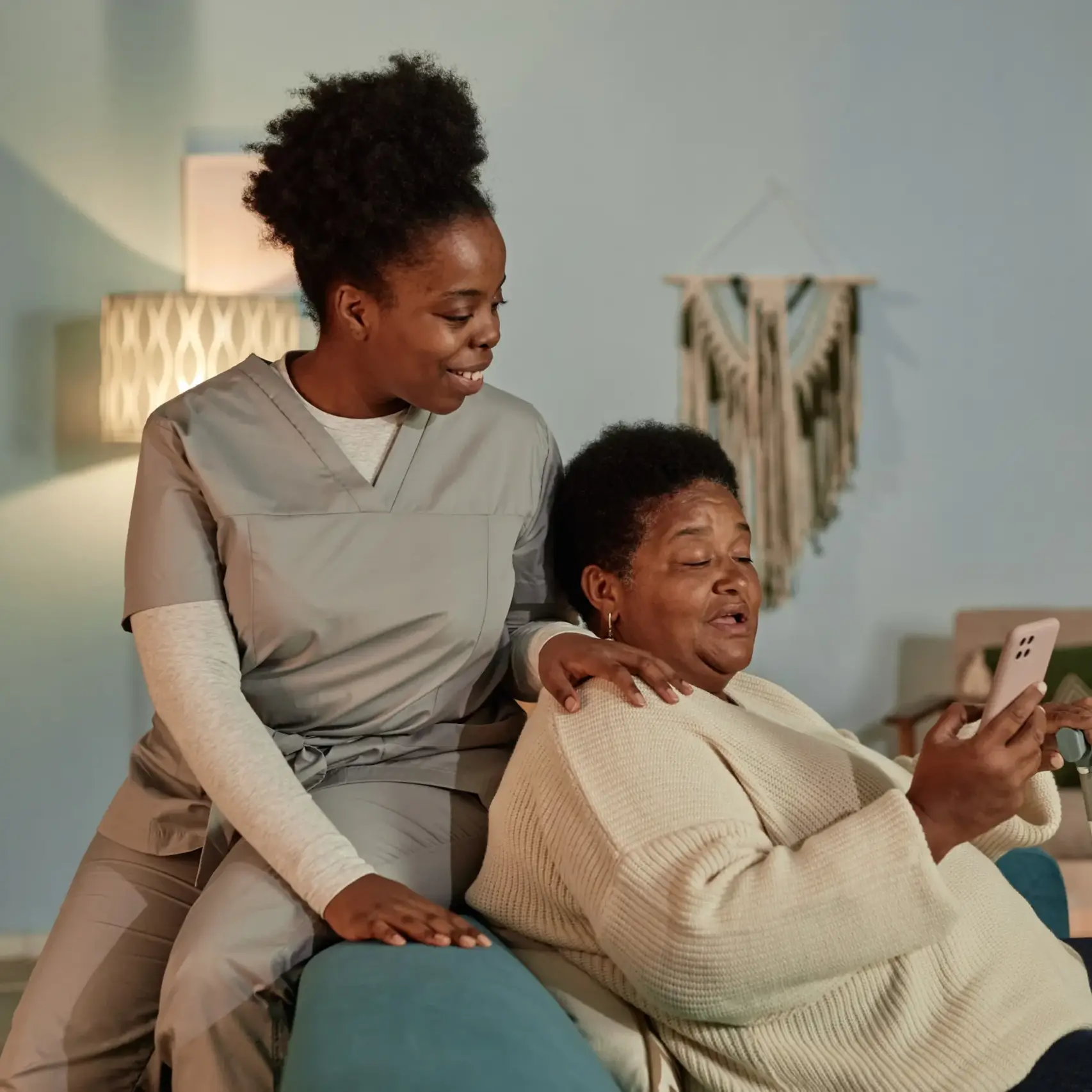 Smiling caregiver supporting elderly woman while she uses a smartphone in a cozy living room.