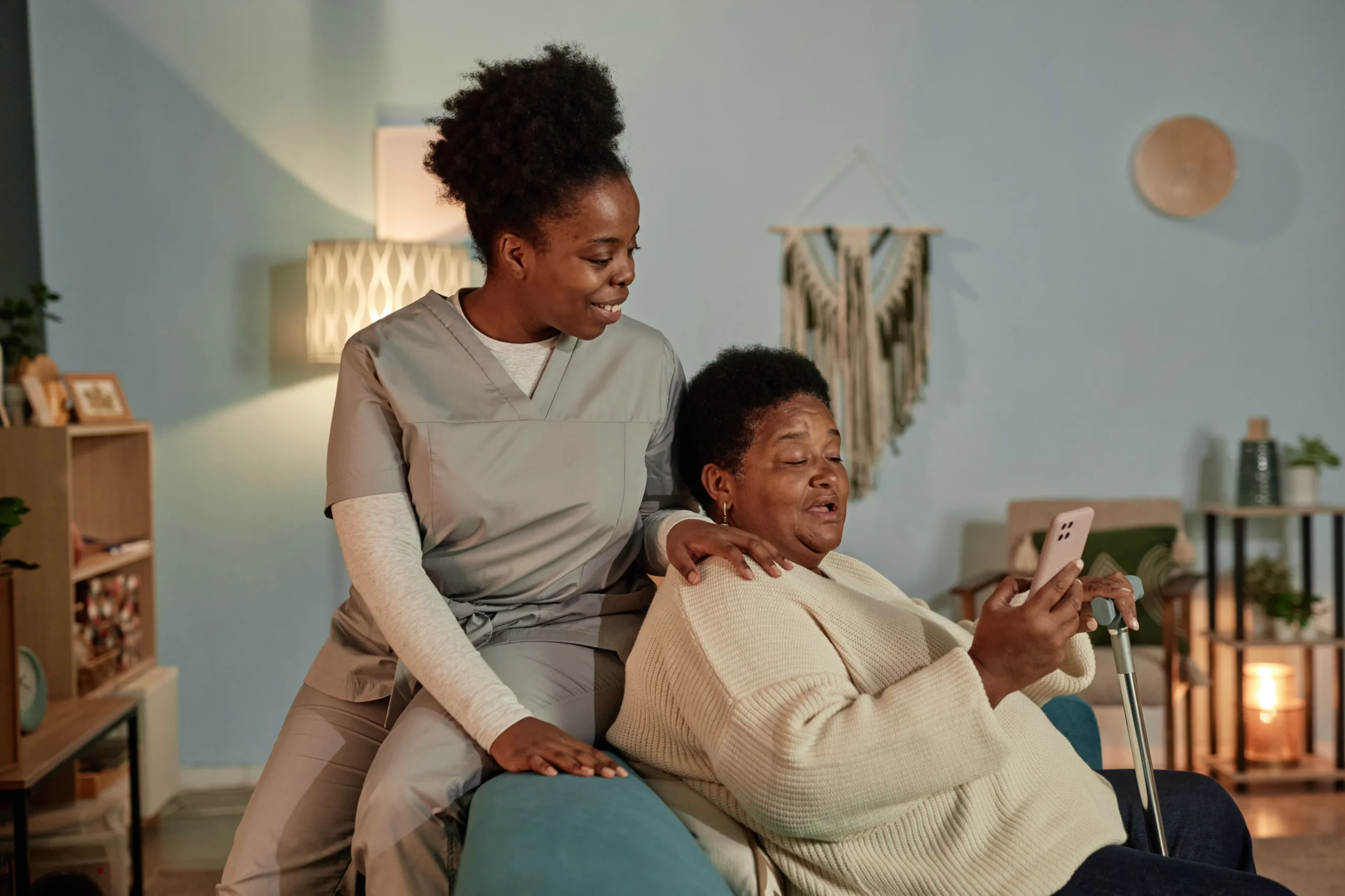 Smiling caregiver supporting elderly woman while she uses a smartphone in a cozy living room.