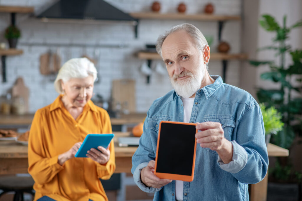 Smiling elderly man showing his tablet while senior woman uses a digital device in the background