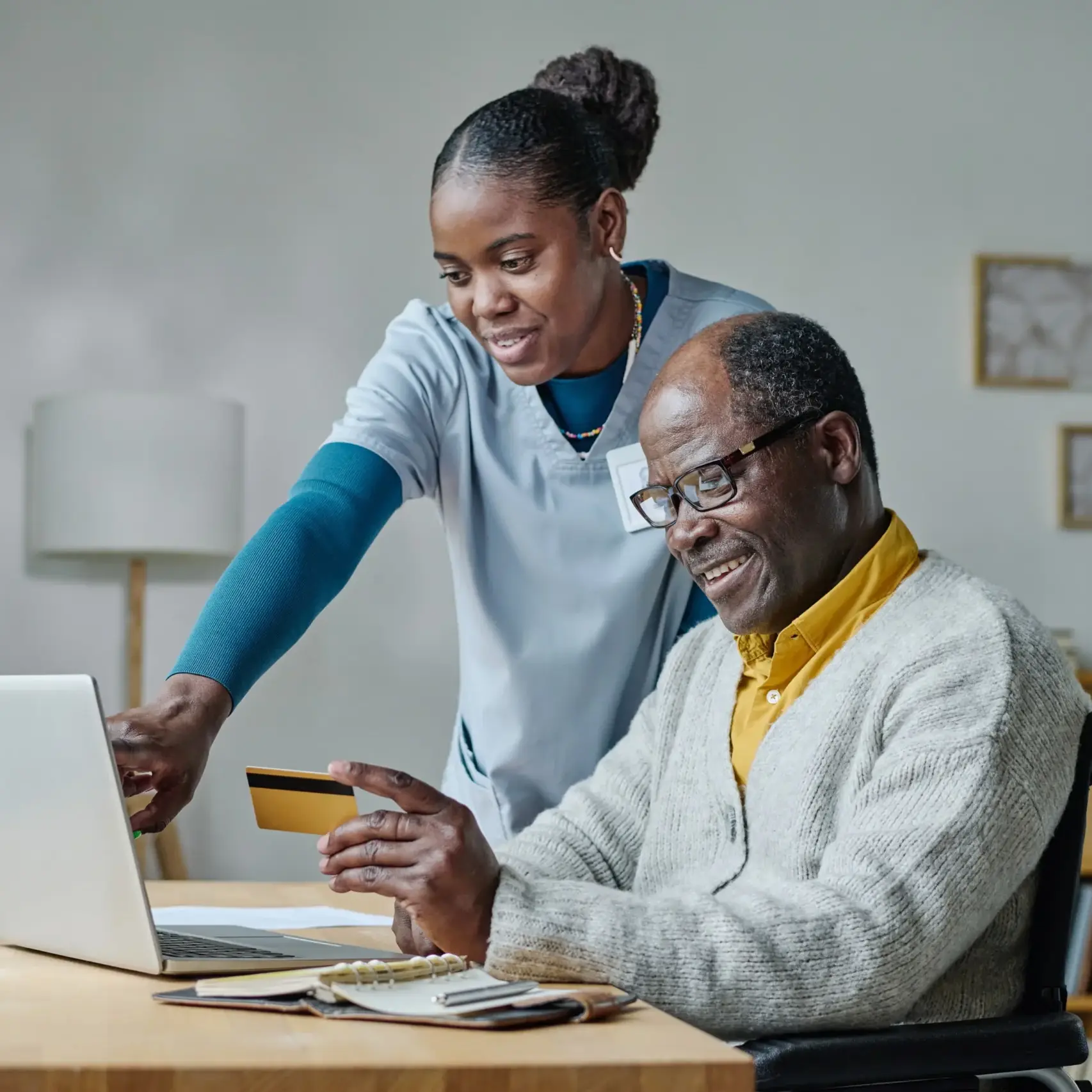 Elderly man in wheelchair using laptop with caregiver assisting online payment