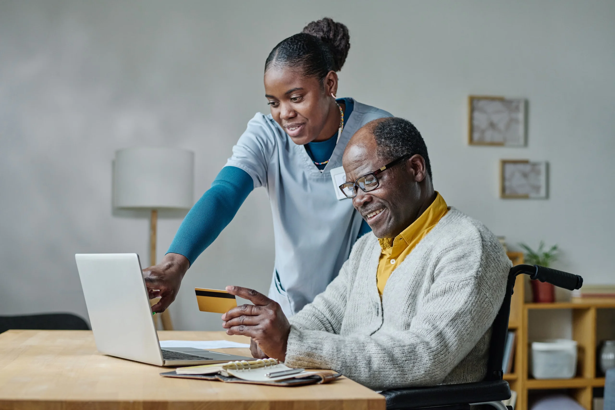 Elderly man in wheelchair using laptop with caregiver assisting online payment