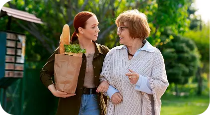 Caregiver walking with an elderly woman outdoors while carrying groceries.