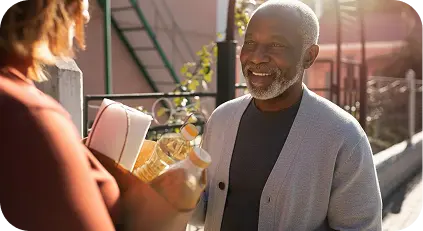 Caregiver handing groceries to an elderly man outdoors.