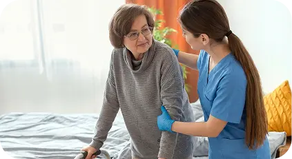 Caregiver in blue scrubs helping an elderly woman with glasses stand up using a walker beside a bed