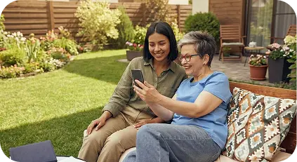 Young woman and older woman with glasses looking at a smartphone together on an outdoor garden bench