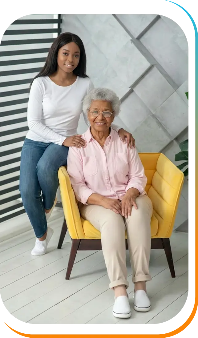 Young woman standing beside an elderly woman with glasses seated in a yellow armchair indoors